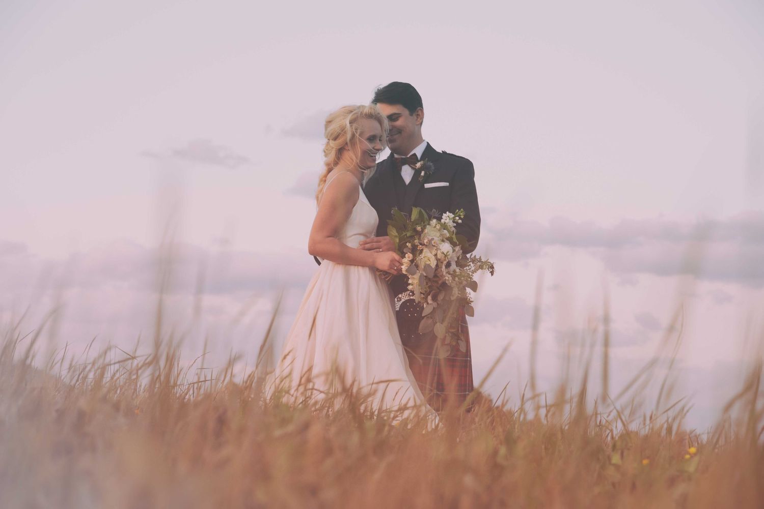 Bride and groom embrace in golden field at sunset, she holds wildflower bouquet in romantic wedding portrait.