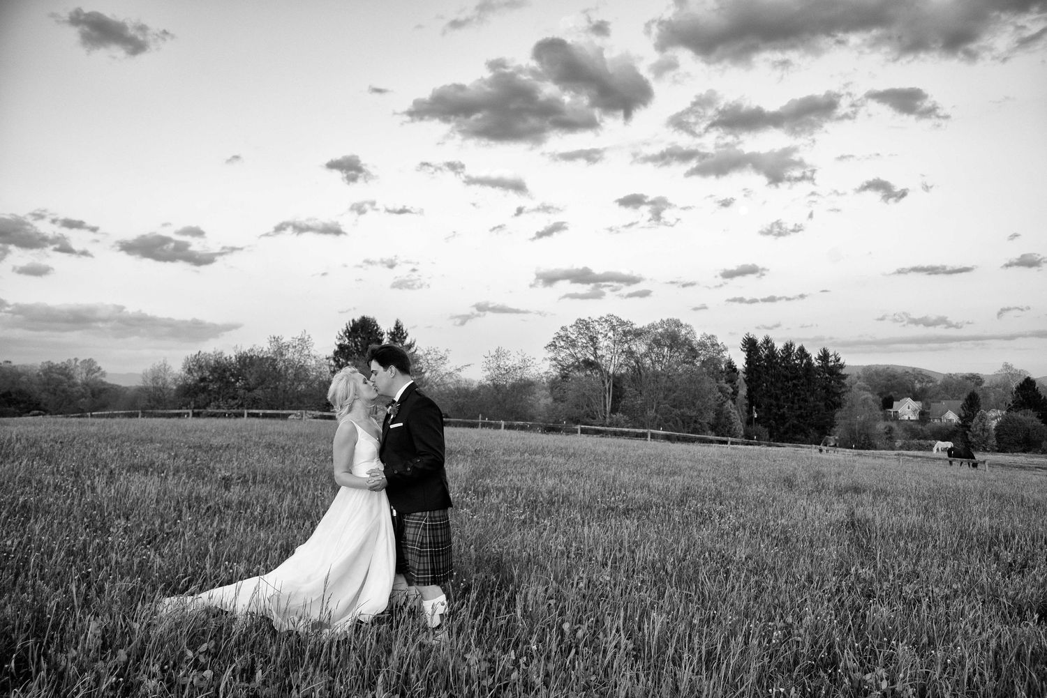 Black and white photo of a bride and groom kissing in a lush green field at sunset on their wedding day.