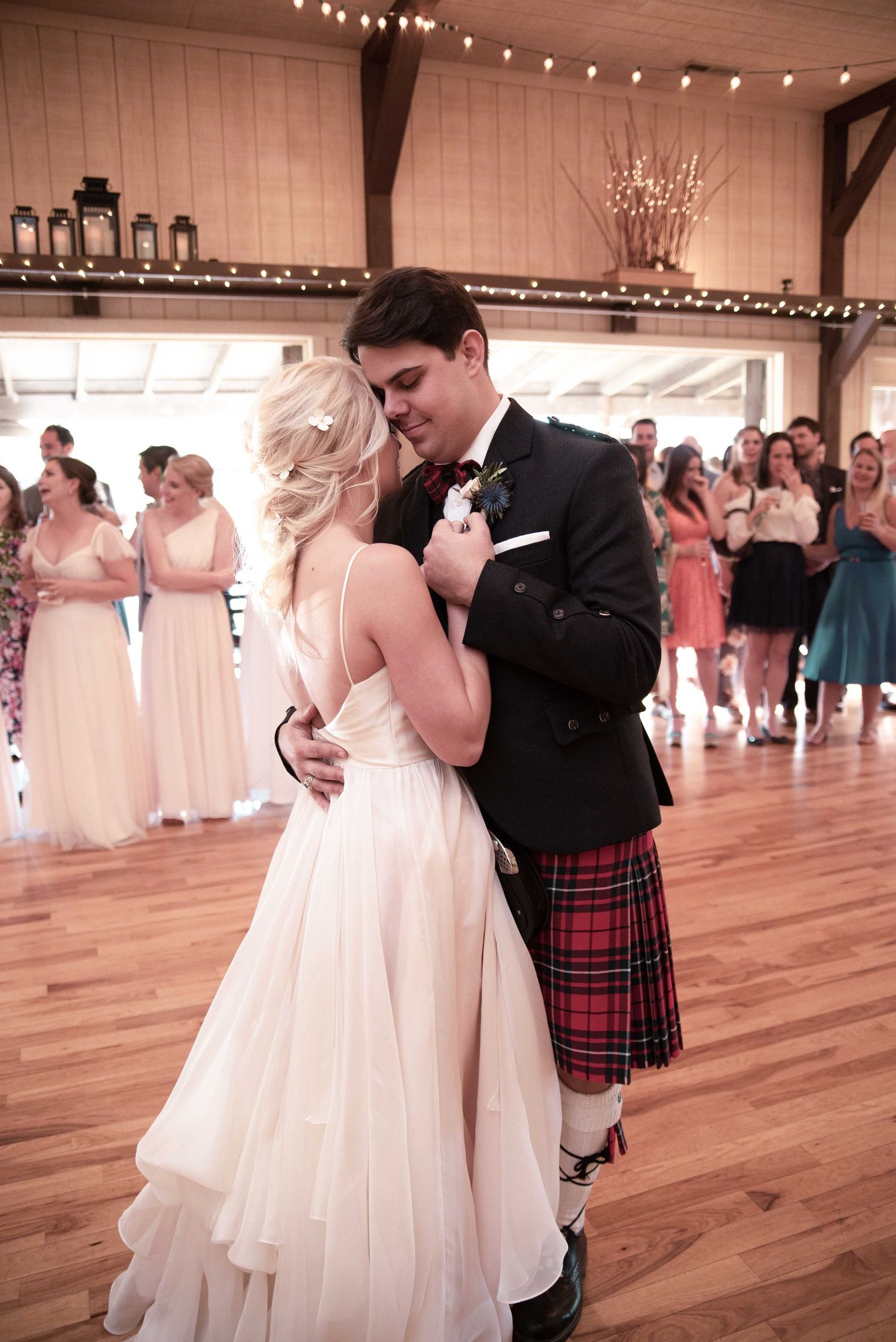 Bride and groom share first dance in rustic barn venue, groom wearing traditional Scottish tartan kilt and black tuxedo jacket.