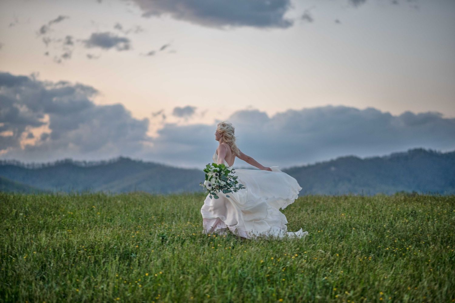 Bride in flowing white gown holding bouquet runs through green meadow with mountain sunset backdrop.
