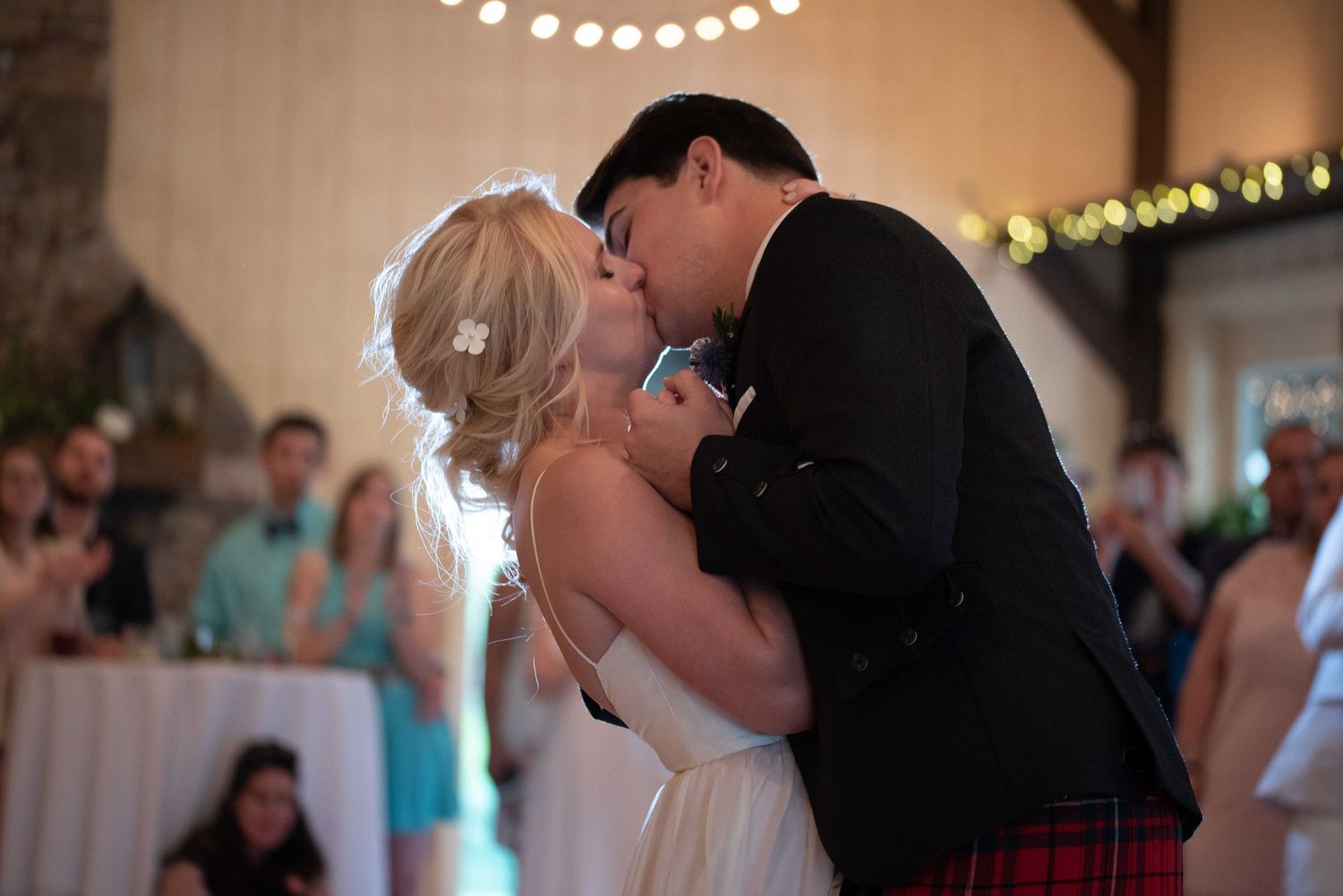 Bride and groom share first dance kiss in rustic barn wedding venue, groom wearing traditional red Scottish kilt.