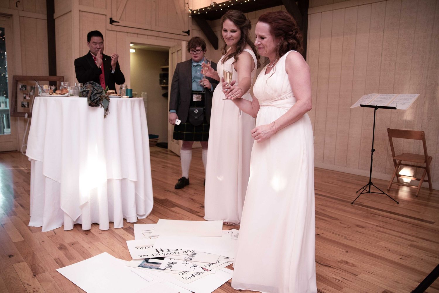 Bridesmaids in white dresses admire wedding artwork on floor at rustic venue reception.