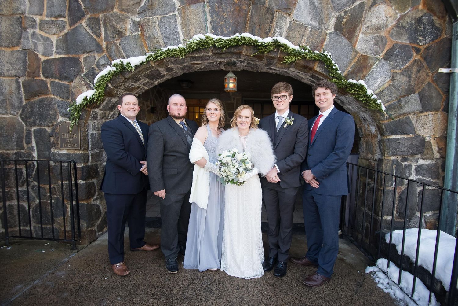 Bride and groom with wedding party standing outside a stone arch venue in winter.