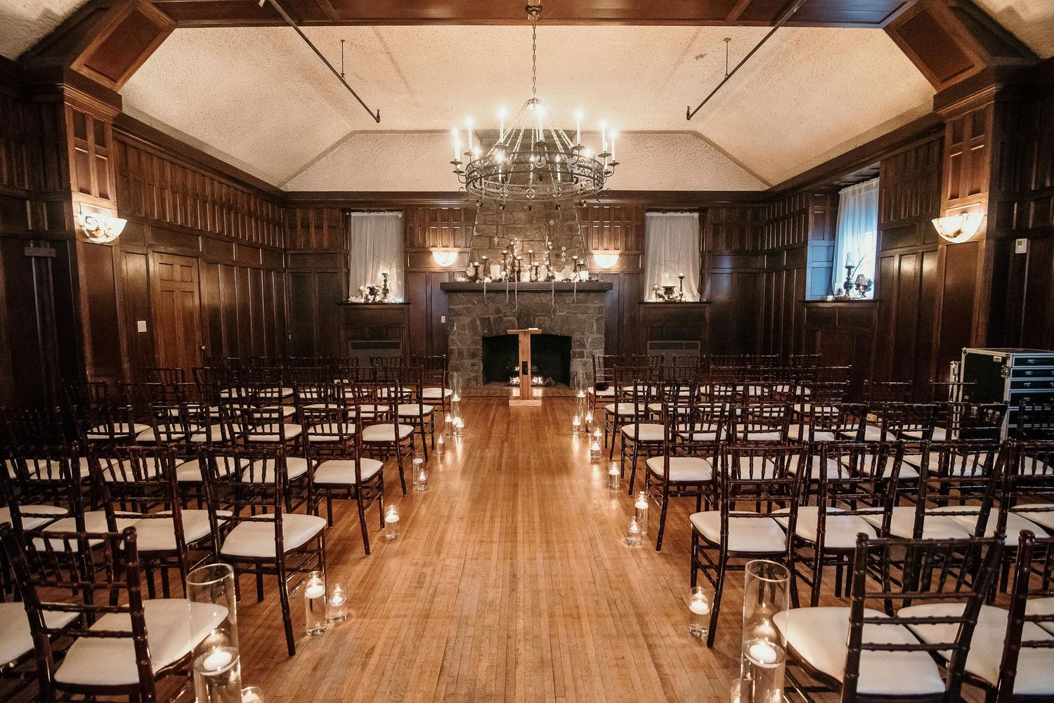Elegant wood-paneled wedding ceremony venue with chiavari chairs, chandelier, stone fireplace, and candlelit aisle on hardwood floors.