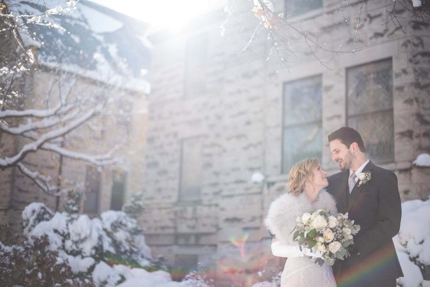 Bride and groom embracing in snowy winter wedding portrait outside stone building with snow-covered trees.