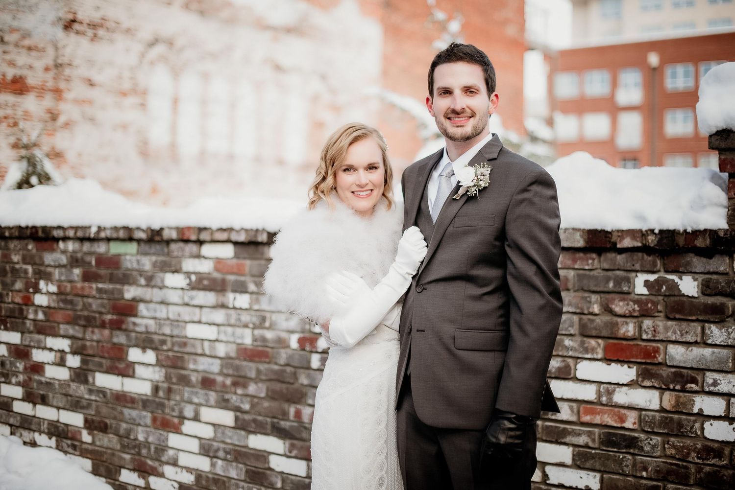 Bride and groom posing together in winter snow beside a brick wall, bride wearing a white fur wrap.