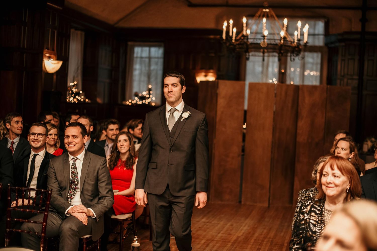 Groom stands at wedding ceremony aisle in gray suit, guests seated in elegant wood-paneled venue with chandelier.