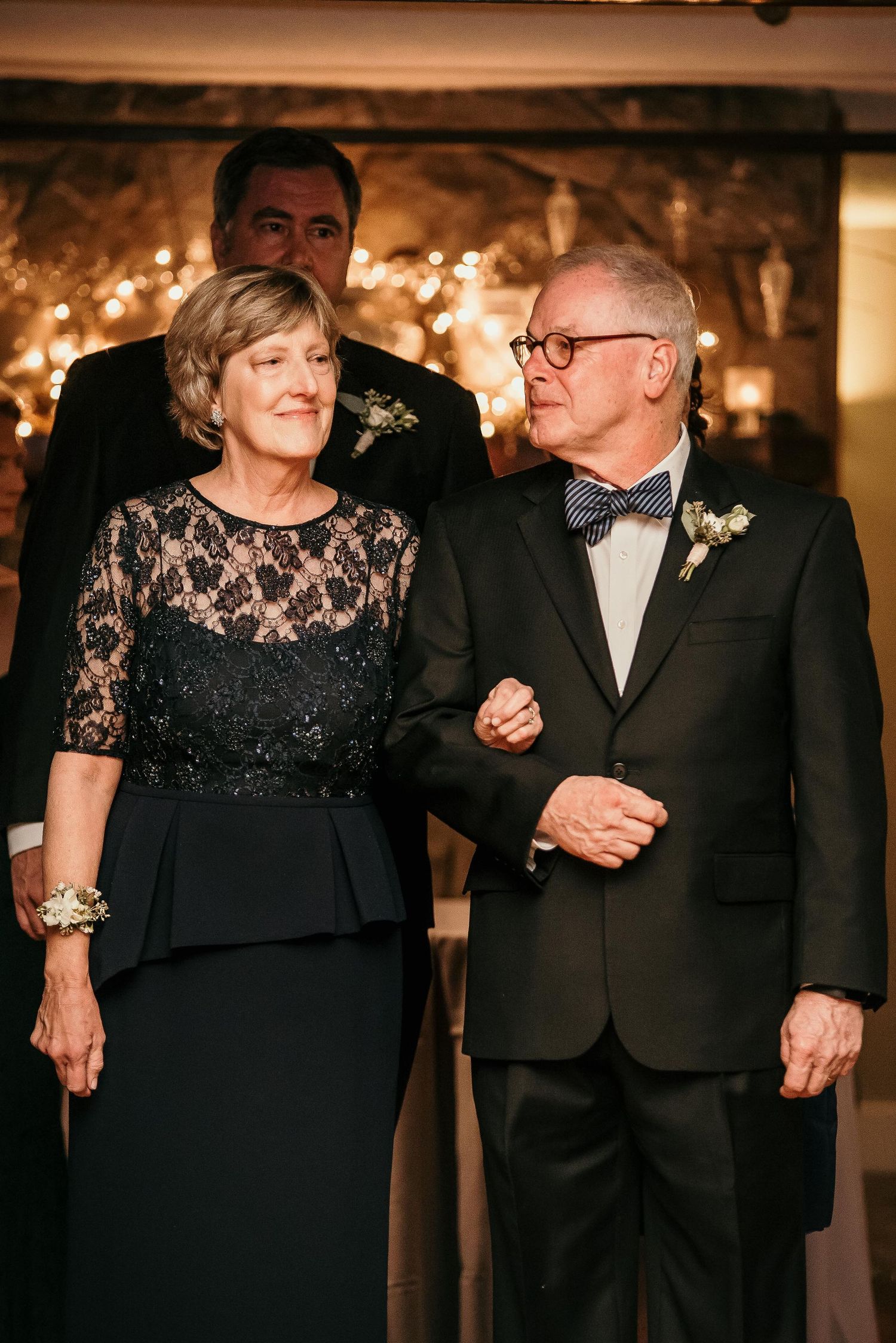 Elegant couple in formal attire at a wedding reception, woman in black lace dress, man in tuxedo with bow tie.