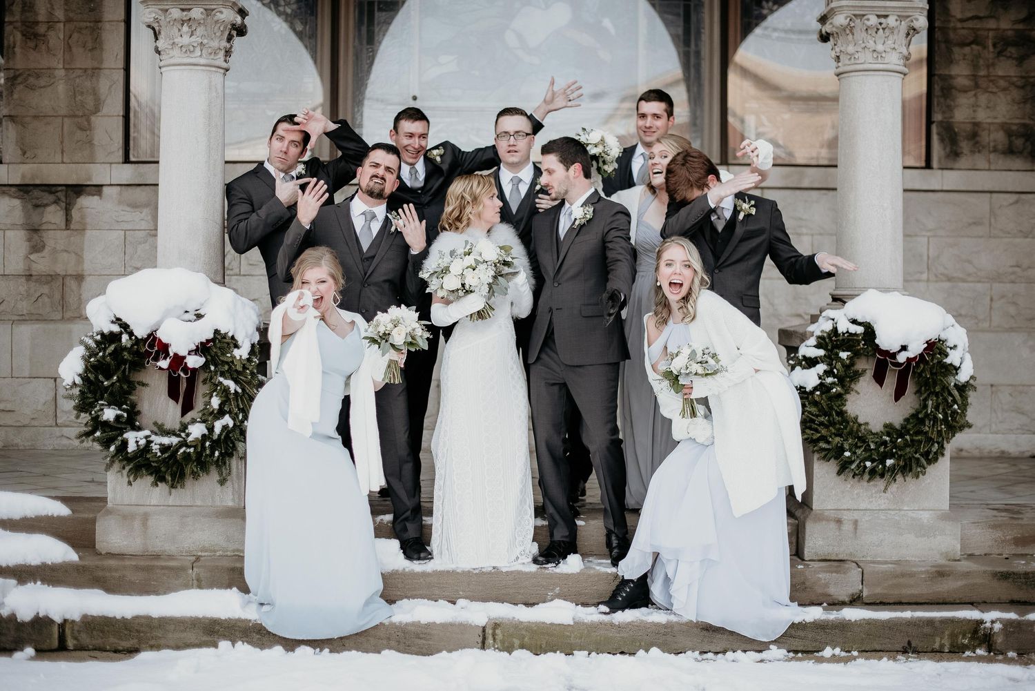 Winter wedding party poses on snowy church steps, bridesmaids in blue, groomsmen in gray suits, bride holding white bouquet.