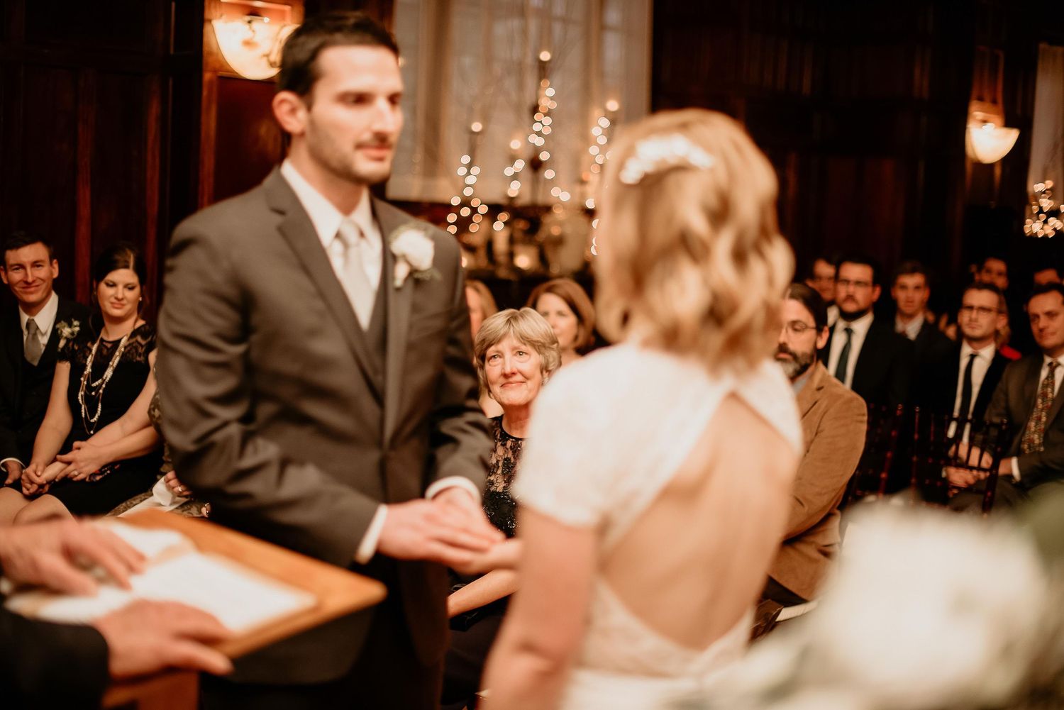 Groom in gray suit holds bride's hands during indoor wedding ceremony with guests seated in background.