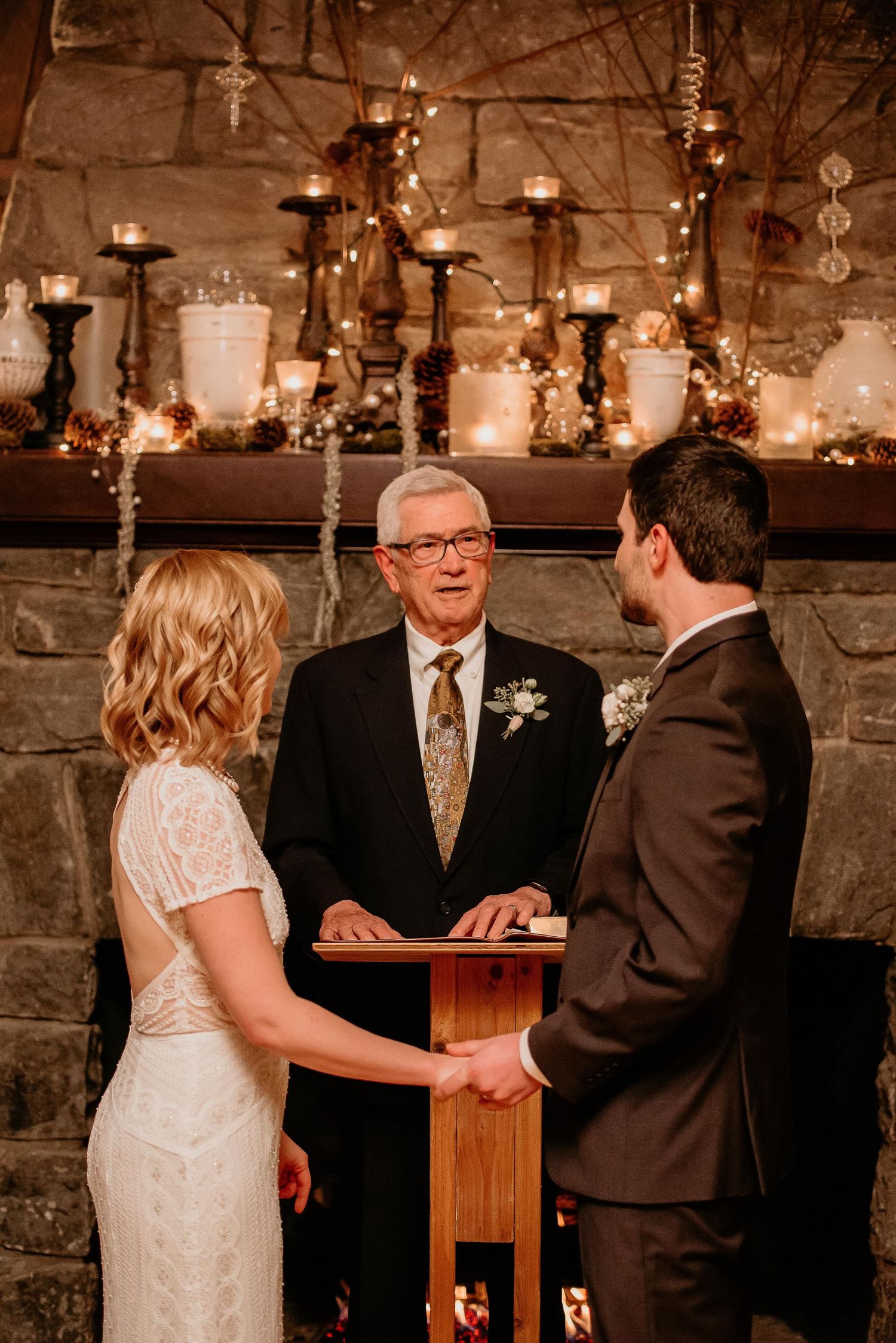 Bride and groom hold hands during wedding ceremony before stone fireplace with candles and officiant at podium.