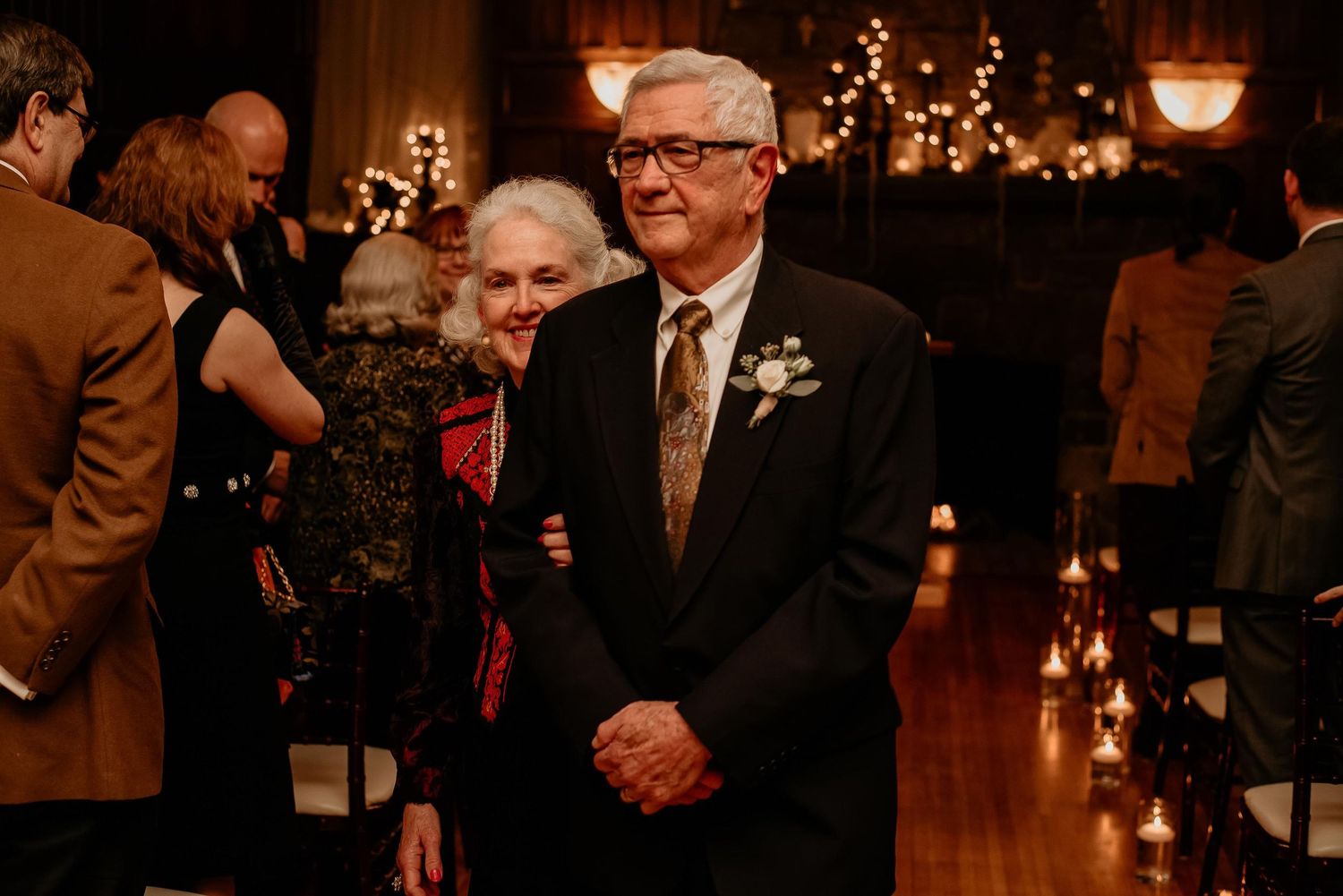 Elegant wedding guests in formal attire stand in a warmly lit venue, man wearing boutonniere smiles at ceremony.