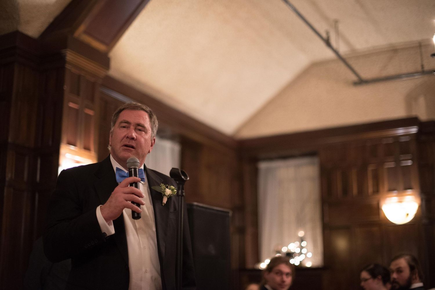 Man in suit speaking at microphone during formal indoor event in wood-paneled room with warm lighting.