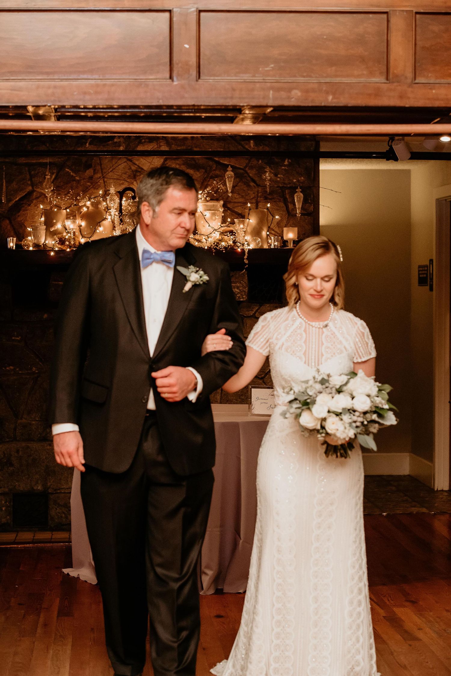 Bride in white lace gown holding bouquet walks arm-in-arm with man in black tuxedo at indoor wedding ceremony.