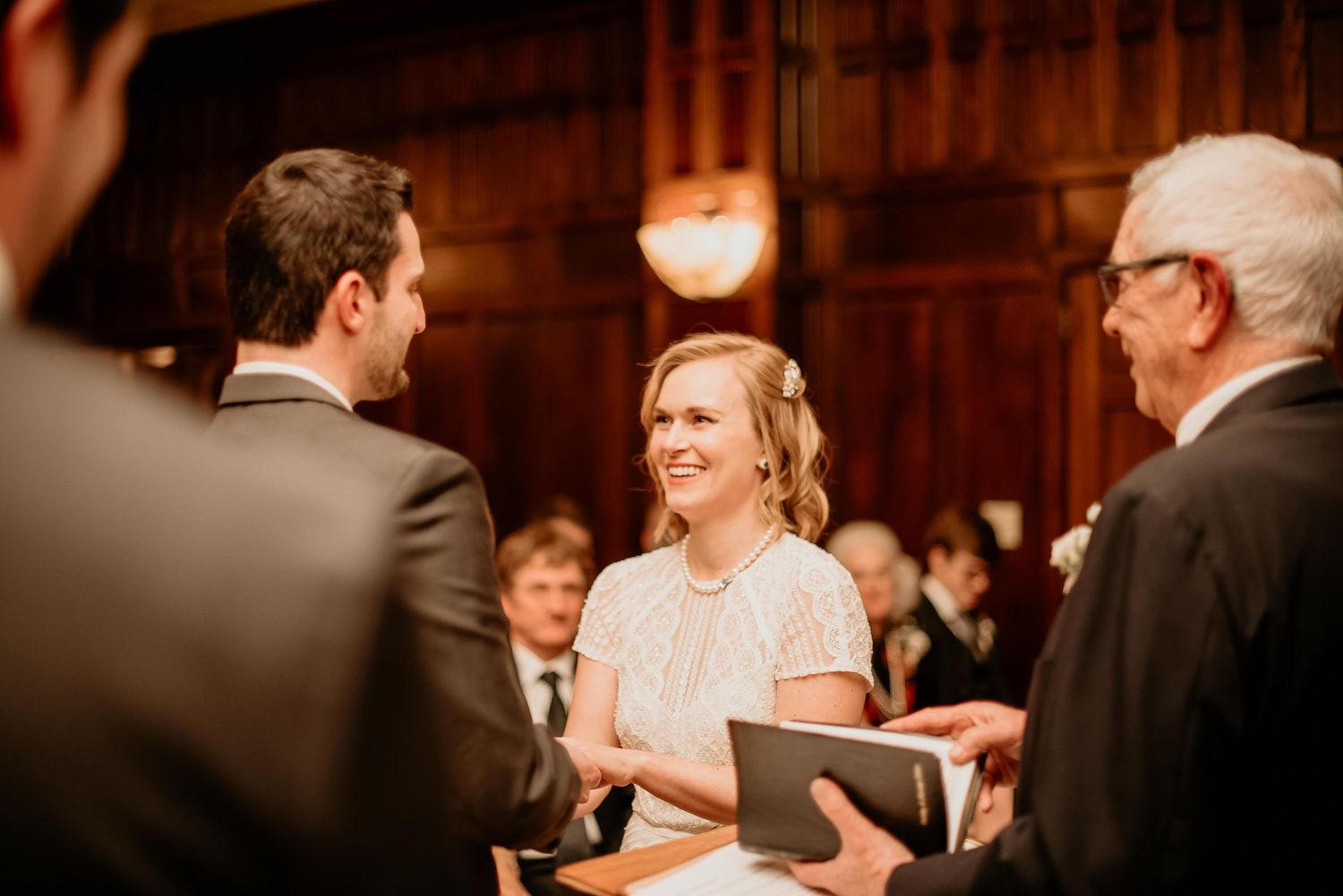 Bride smiling joyfully during wedding ceremony, exchanging vows with groom in elegant wood-paneled venue.