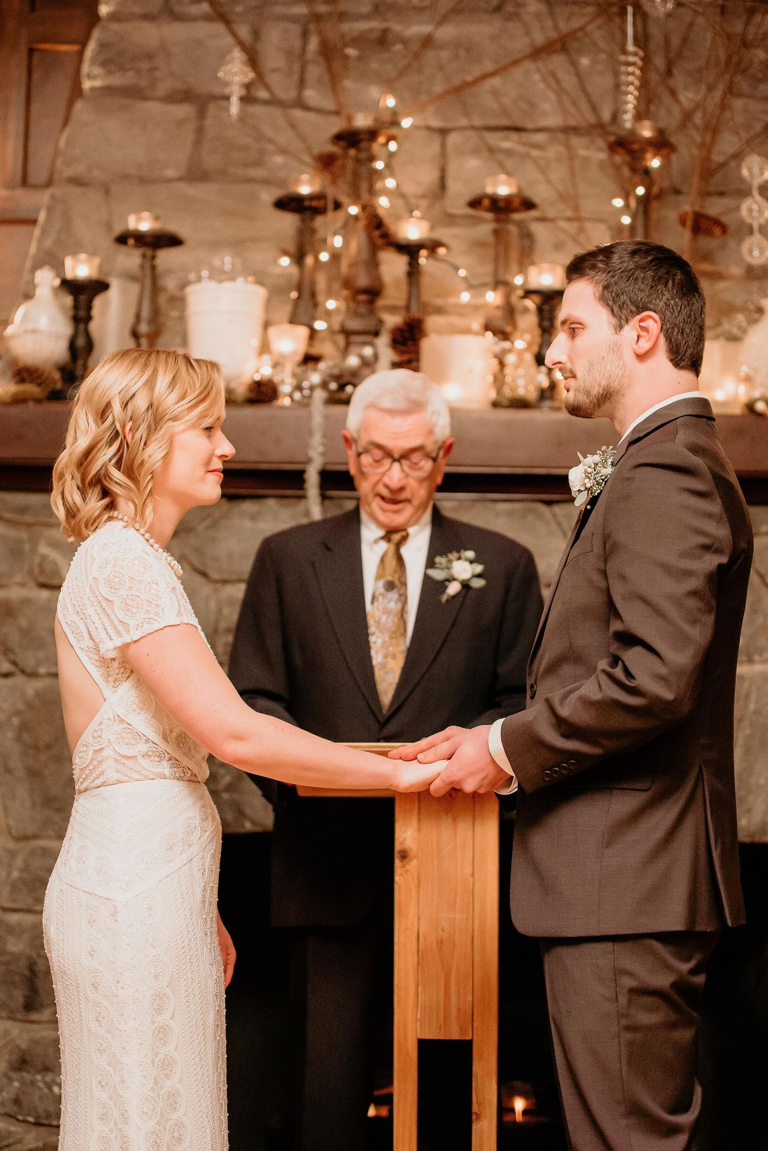 Bride and groom exchange vows at rustic stone fireplace altar with officiant and candlelit decor.