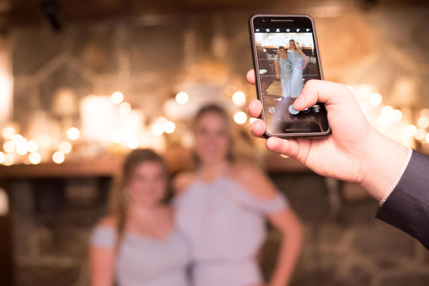 Person photographing two women in gray dresses at a warmly lit wedding reception venue.