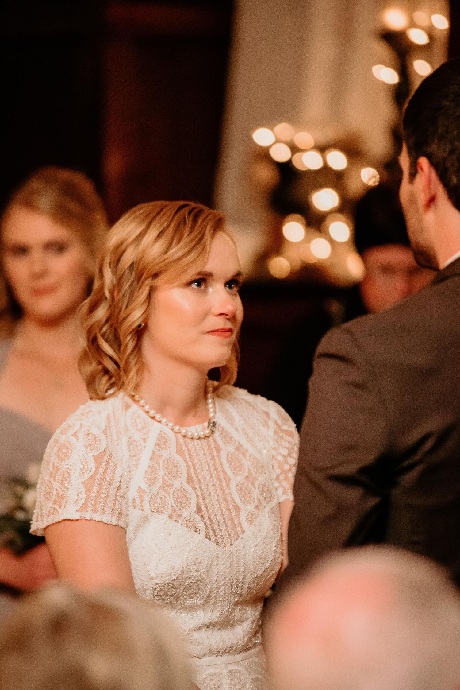 Bride in lace dress and pearl necklace gazes at groom during wedding ceremony with warm bokeh lights.