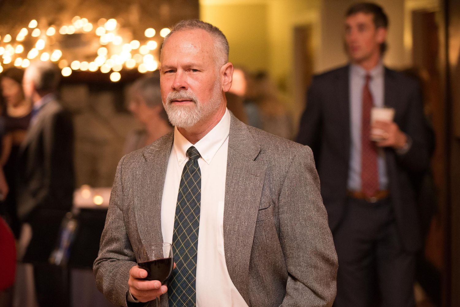 Bearded man in gray suit and dark tie holding a drink at an elegant indoor event with warm lighting.