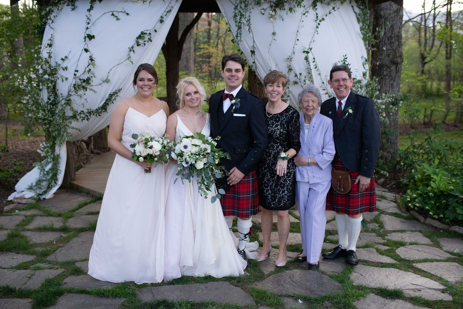 Wedding party posing outdoors under floral arch, bride in white, groom in tuxedo, man in red tartan kilt.