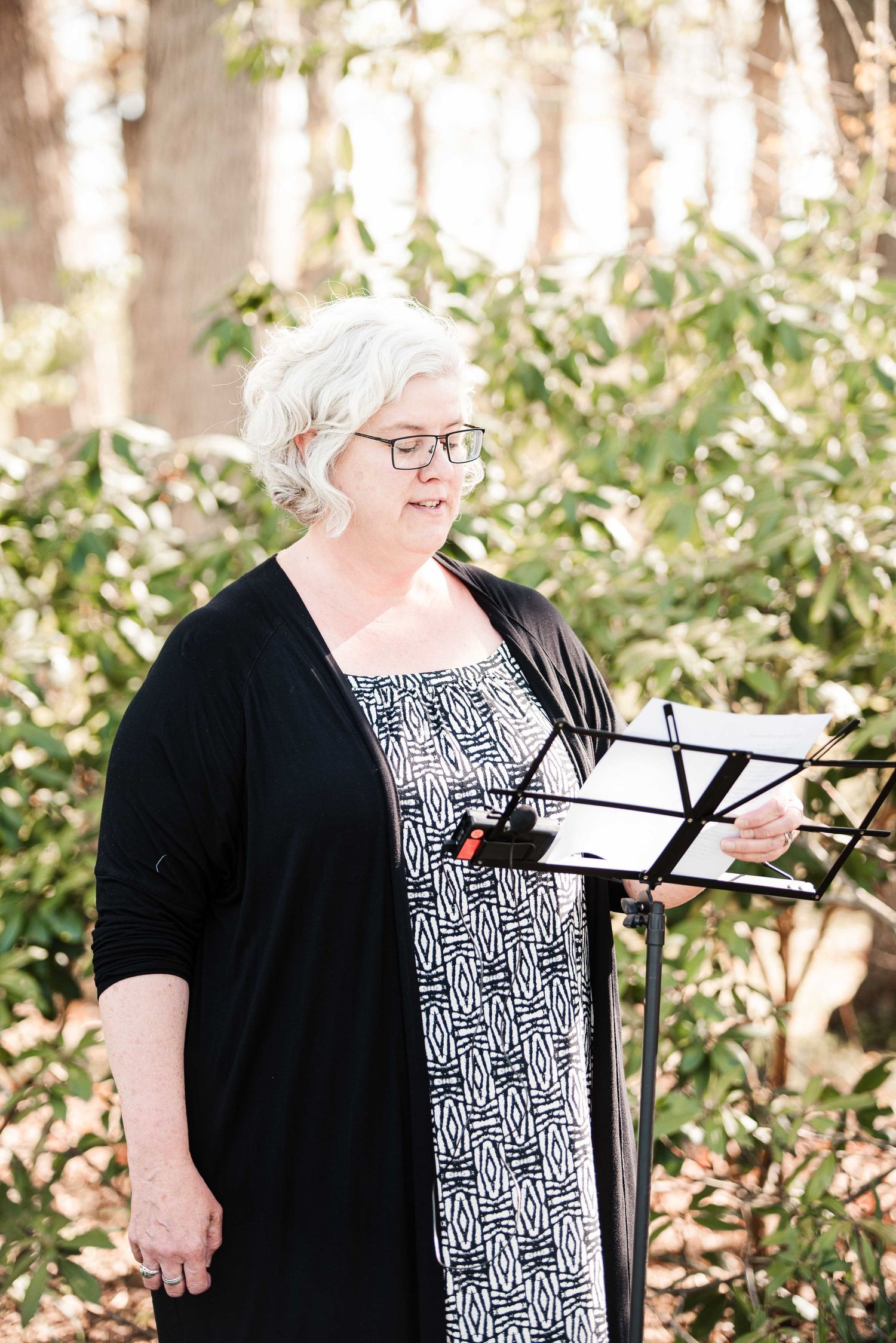 Woman with gray hair and glasses reads from music stand outdoors among green foliage.