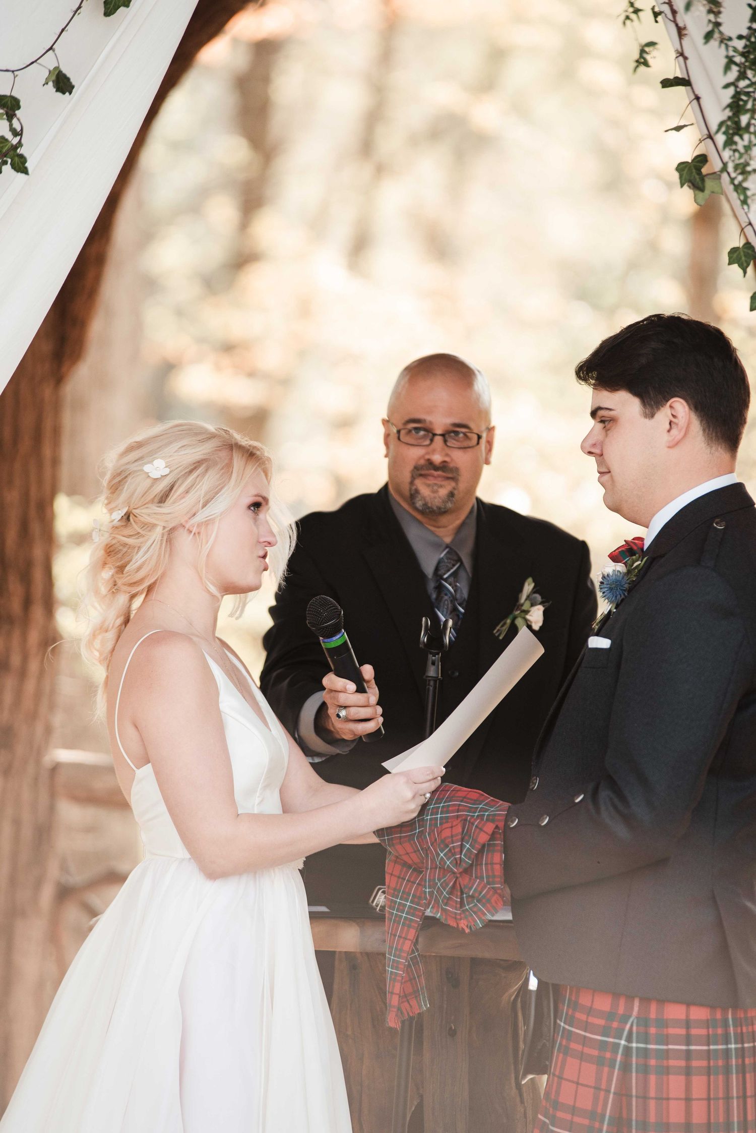 Bride reads wedding vows to groom in Scottish kilt at outdoor ceremony, officiant standing behind them.