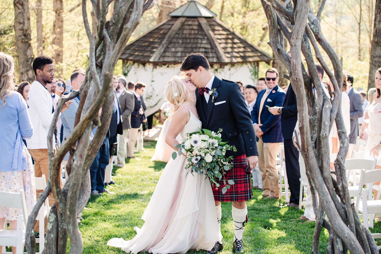 Bride and groom share first kiss at outdoor wedding ceremony under rustic wooden arch, groom wearing traditional Scottish kilt.