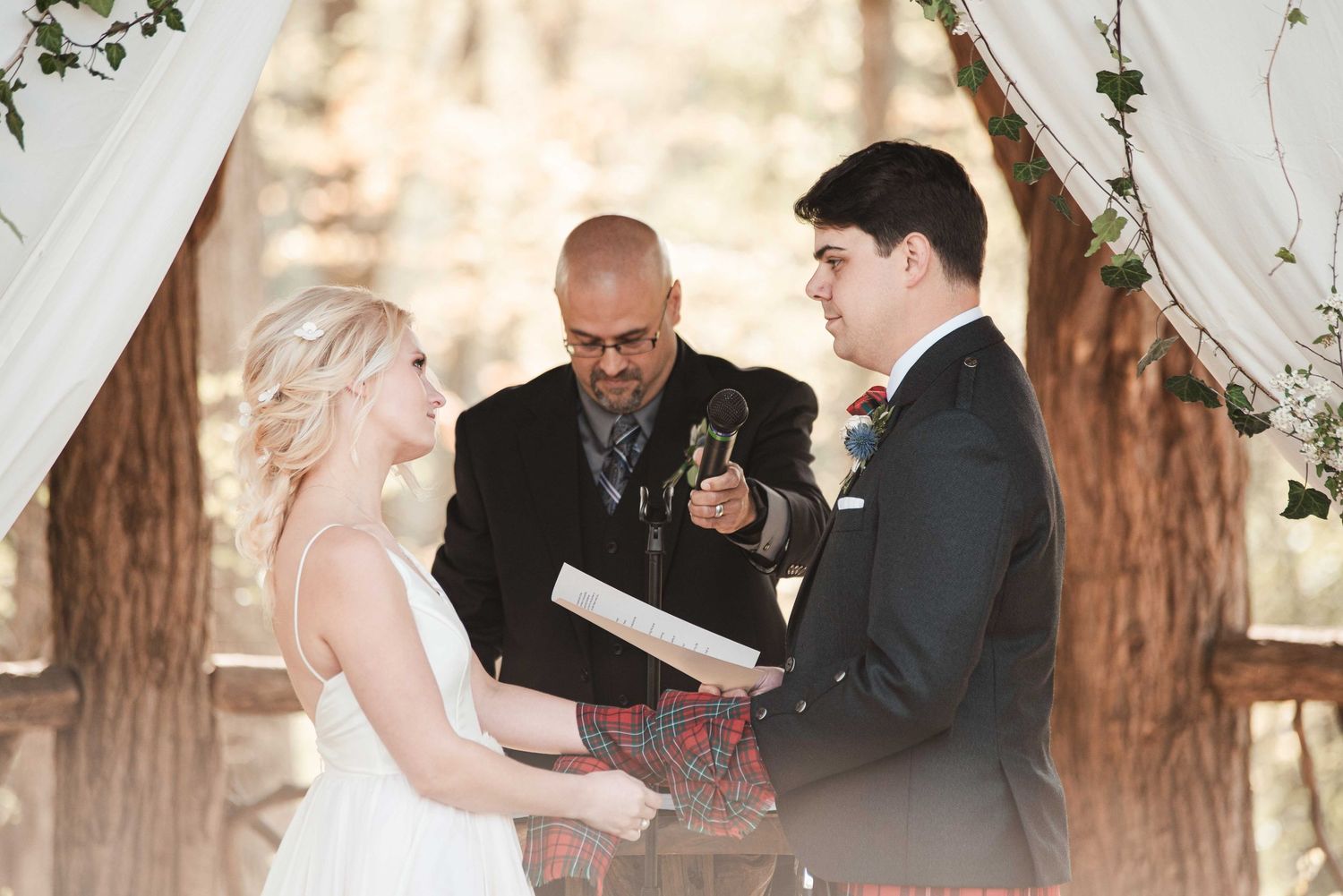 Bride and groom exchange vows with officiant at outdoor woodland wedding ceremony beneath floral arch.