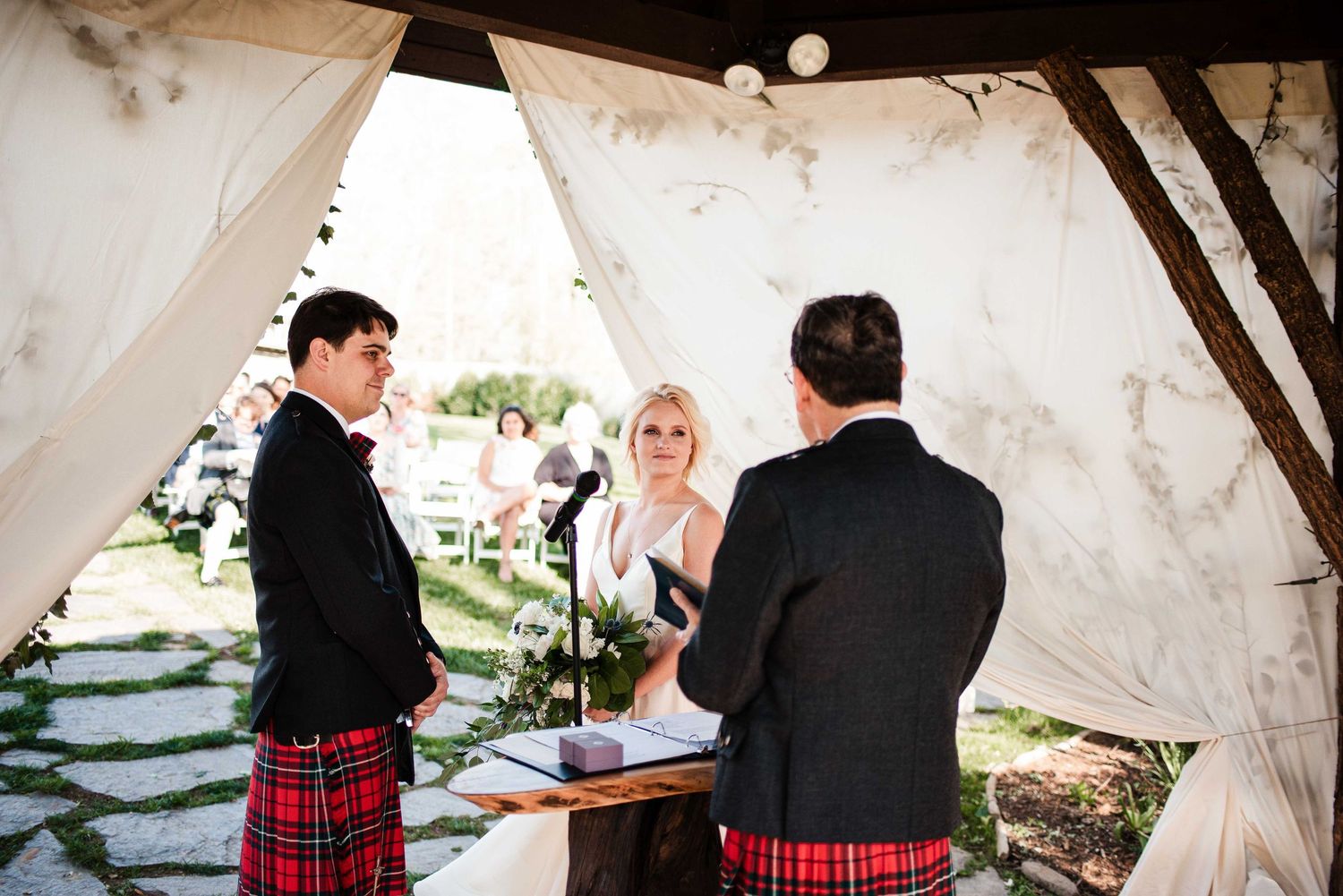 Outdoor wedding ceremony with groom and officiant in red kilts, bride in white dress, under a draped canopy.