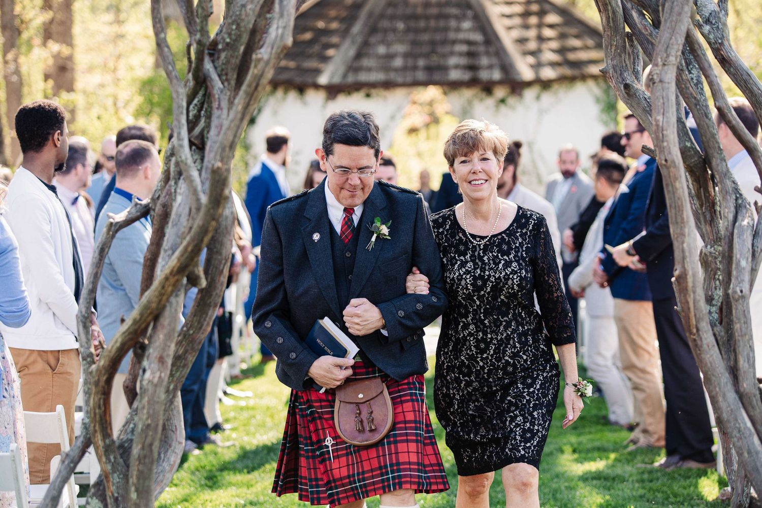Man in Scottish kilt and woman in black dress walk down outdoor wedding aisle lined with rustic wooden trees.