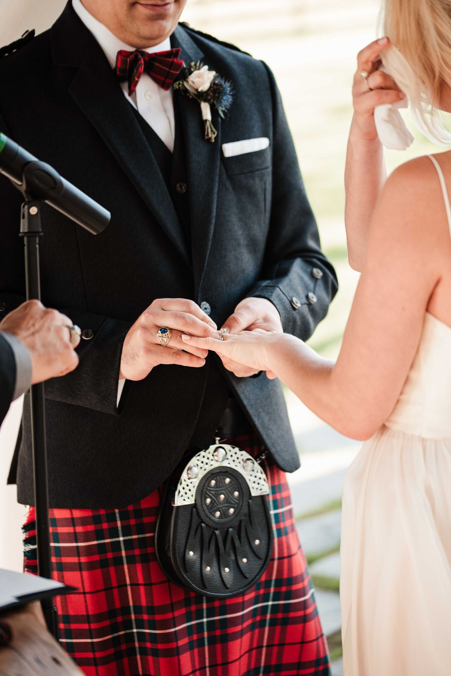 Groom in red tartan kilt exchanges wedding rings with bride during outdoor Scottish wedding ceremony.