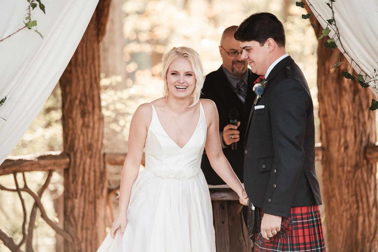 Bride in white gown and groom in red tartan kilt hold hands during outdoor wedding ceremony under trees.