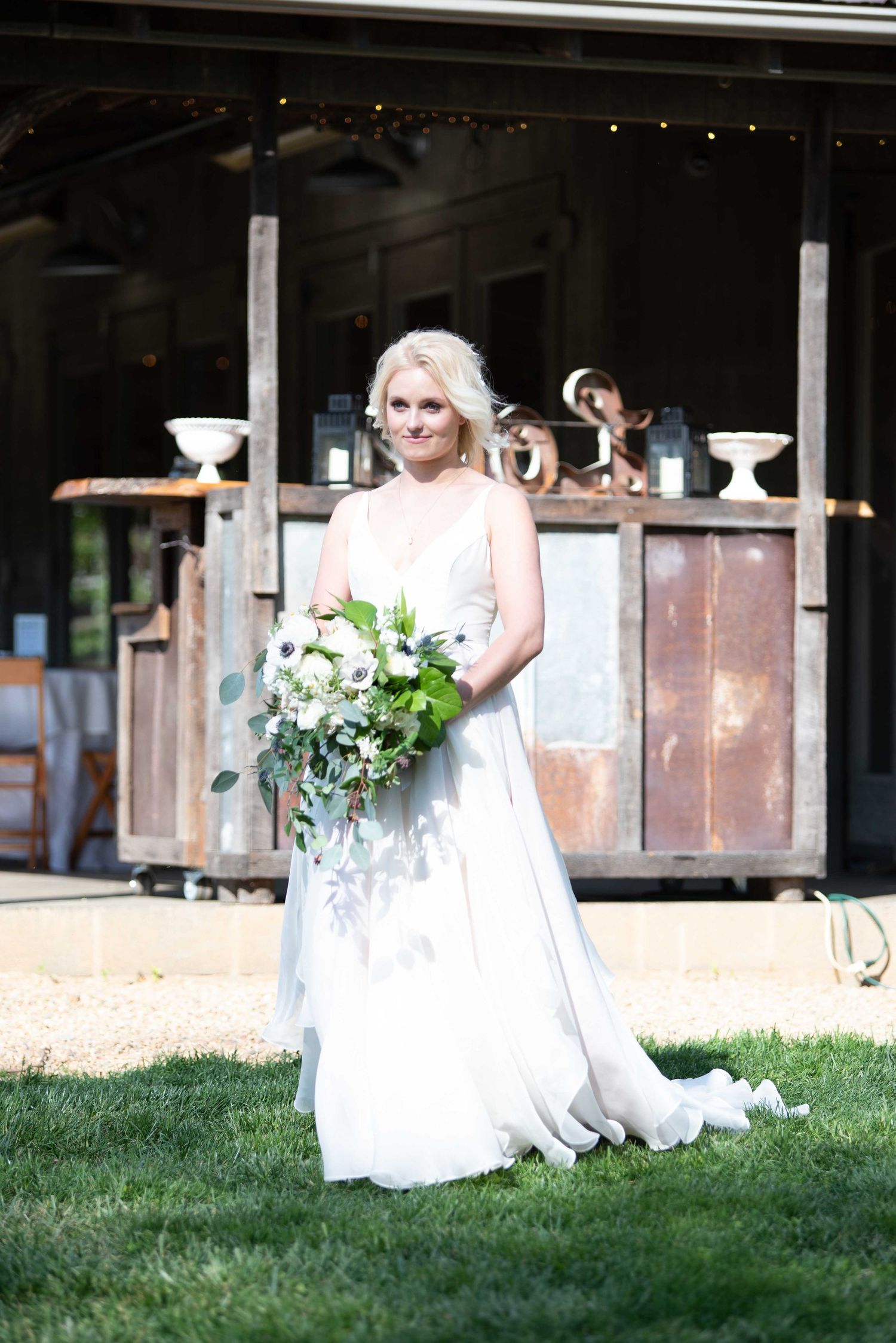 Bride in white gown holding green and white bouquet stands on grass before rustic wooden backdrop.