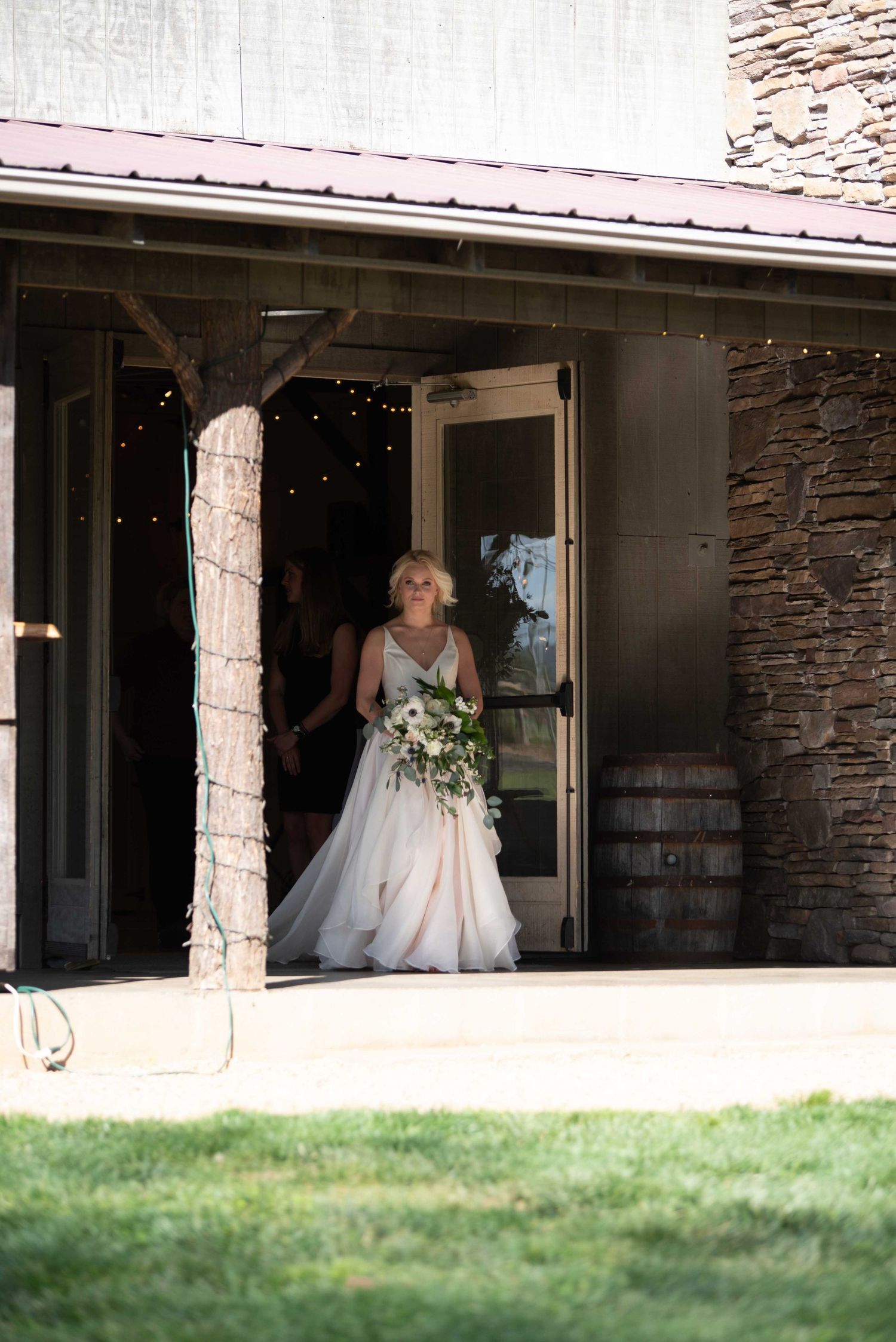 Bride in white gown holding bouquet exits rustic barn venue with string lights and stone walls.