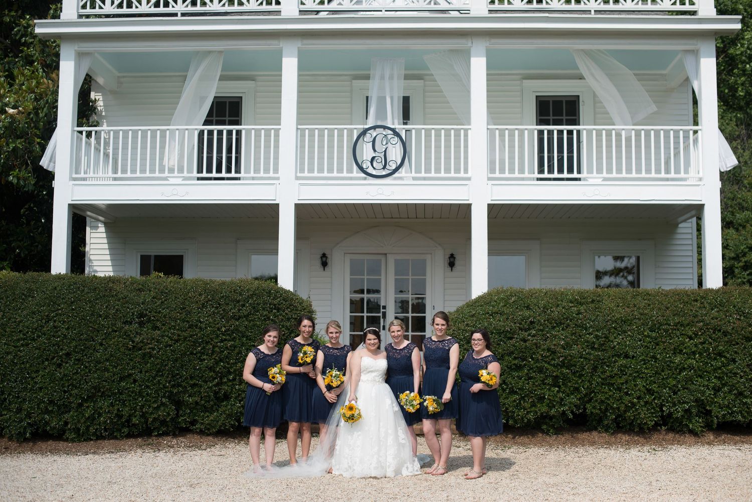 Bride with bridesmaids in navy dresses holding yellow sunflowers posing in front of a white Southern estate.