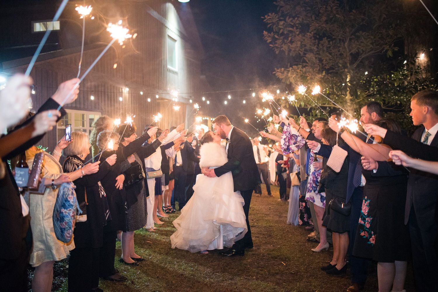 Bride and groom kiss under sparklers held by wedding guests at night.