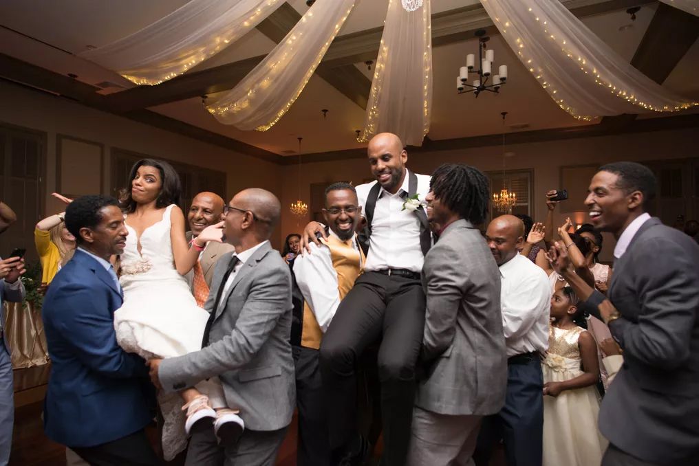 Joyful wedding reception as groomsmen lift the bride and groom in celebration inside an elegant decorated ballroom.