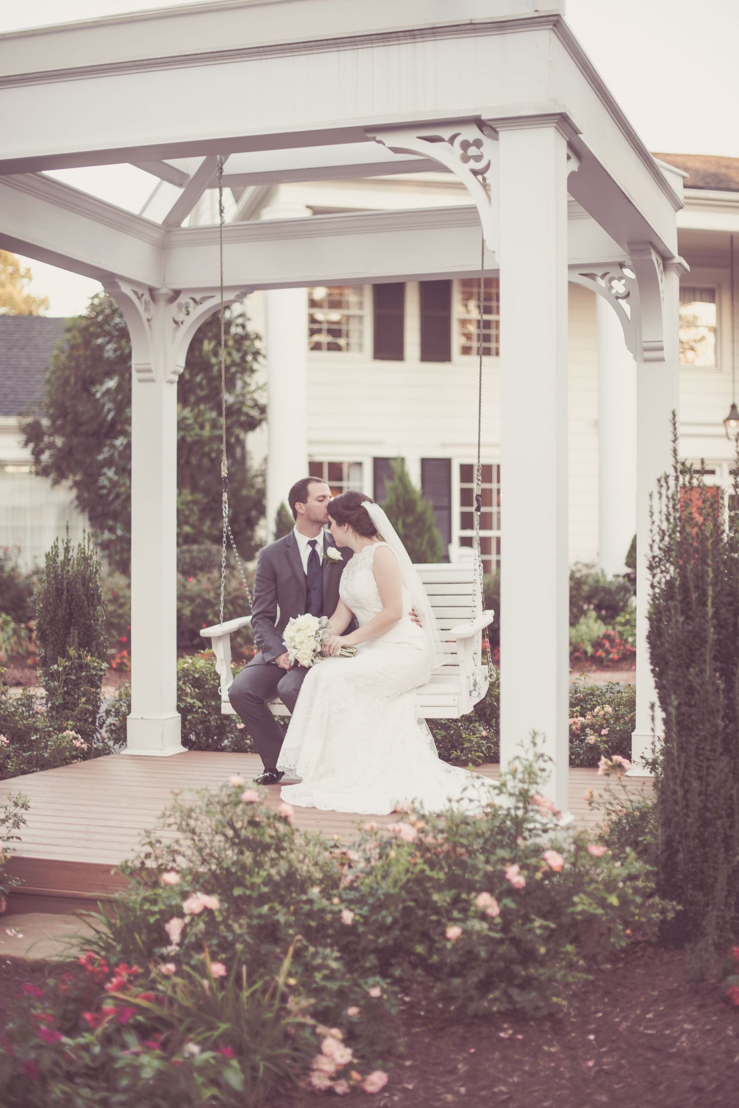 Bride and groom sharing a kiss on a white porch swing surrounded by garden roses.