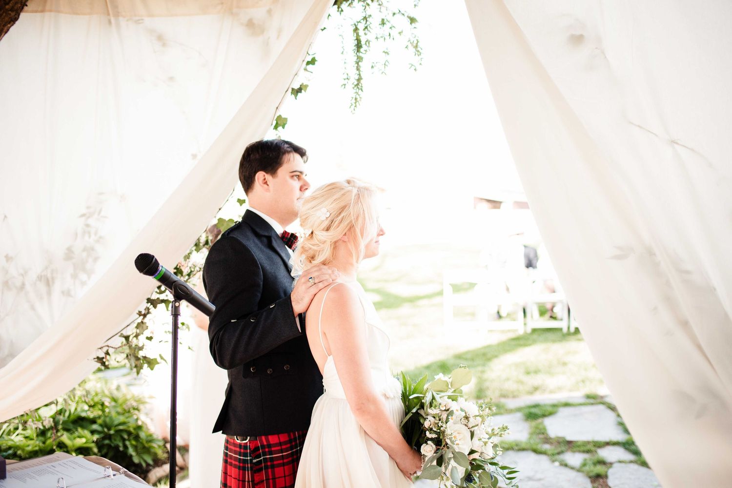 Bride and groom share a romantic moment at outdoor wedding ceremony under white draped canopy with greenery.