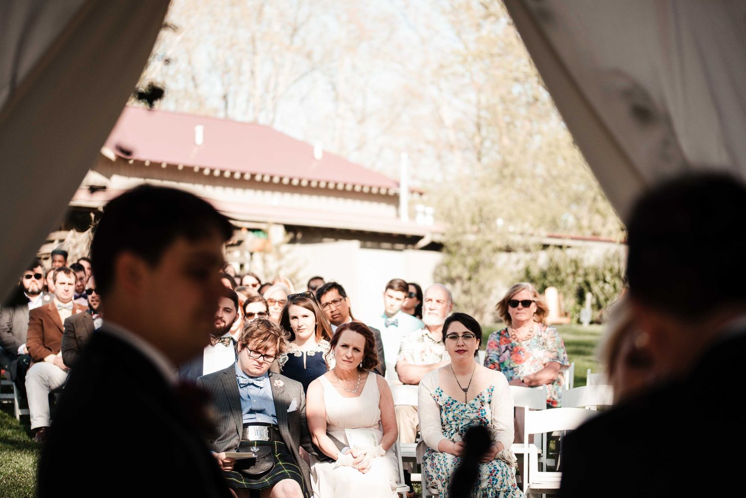 Guests seated outdoors at a wedding ceremony, smiling and watching the proceedings on a sunny day.