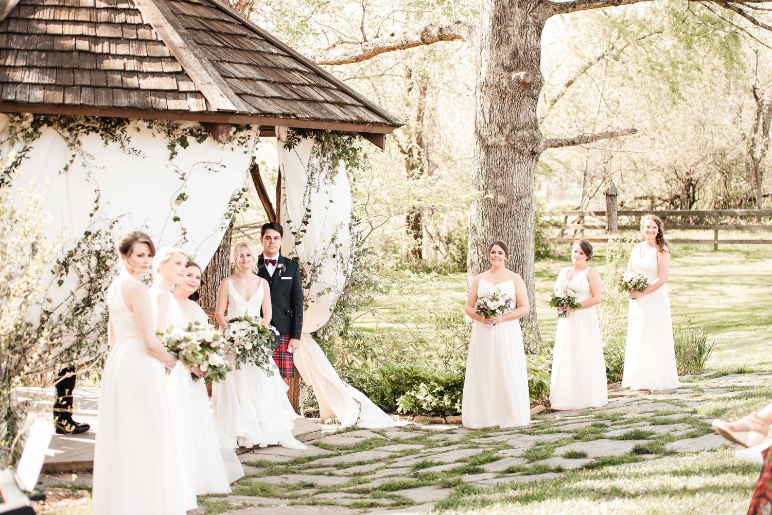 Bridal party in white gowns poses near a floral arch gazebo on a sunny day at an outdoor wedding venue.