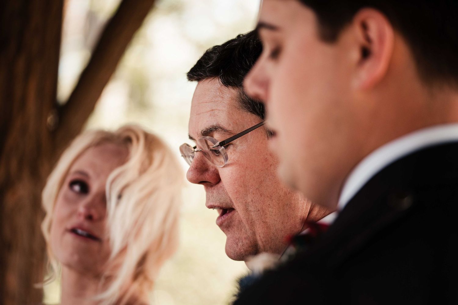 Three people in formal attire at an outdoor event, man with glasses in profile view flanked by blonde woman and young man.