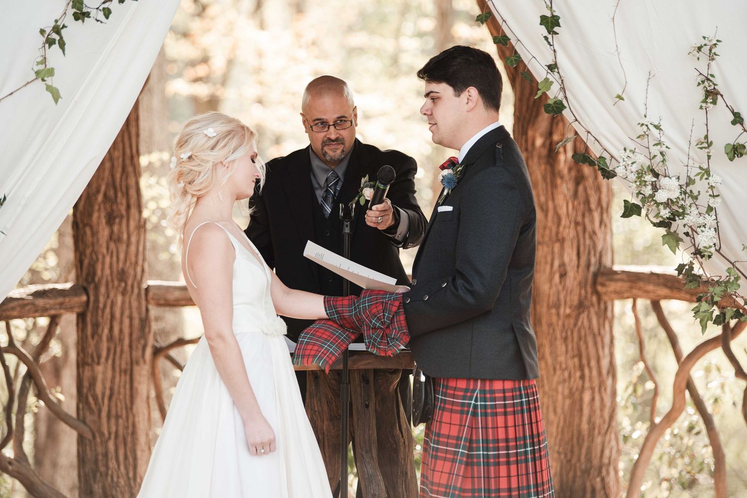 Bride and groom exchanging vows at rustic woodland altar with officiant in tartan kilt.