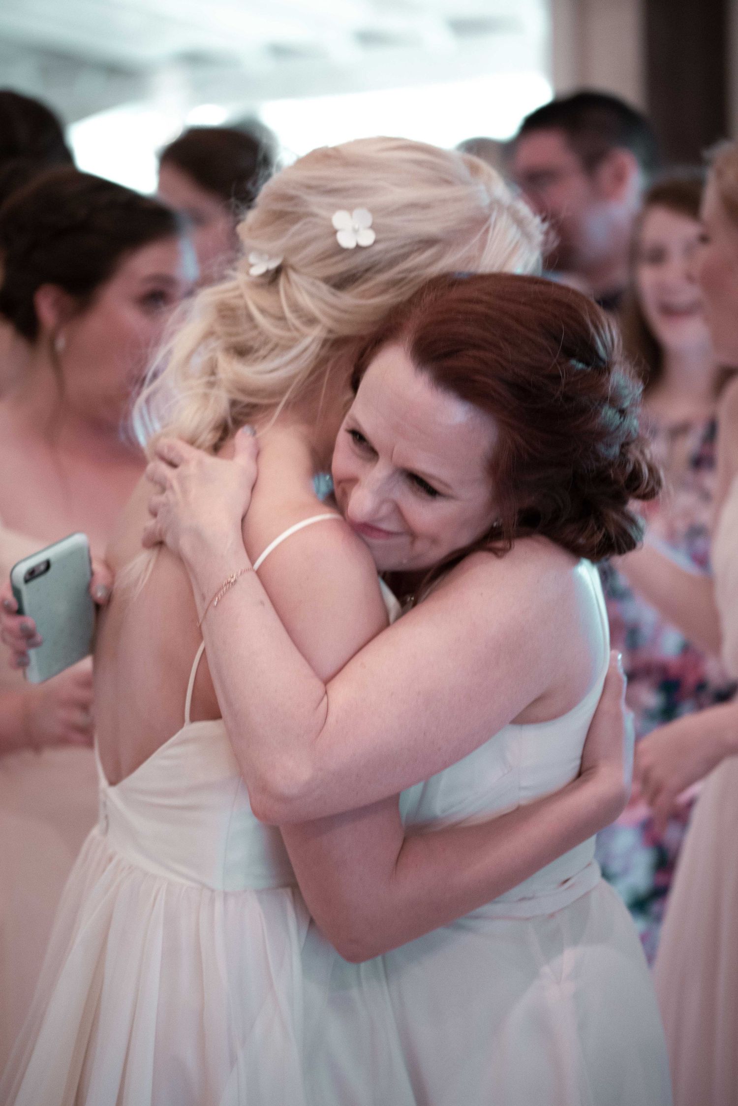 Two women in white dresses share an emotional hug at a wedding celebration surrounded by guests.
