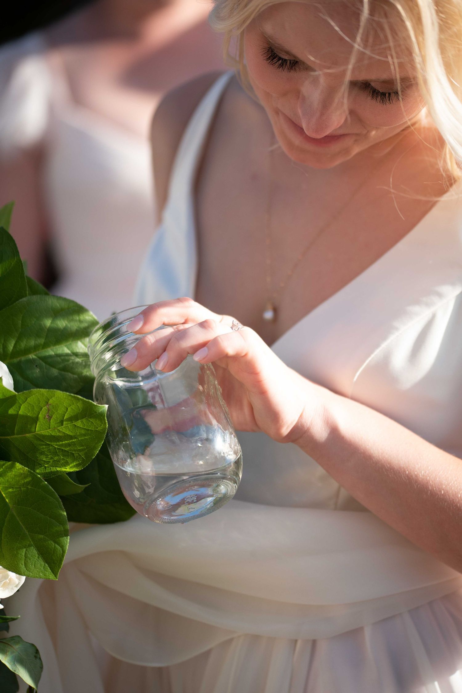 Blonde woman in white dress holding a glass jar with green leaves in soft natural light.