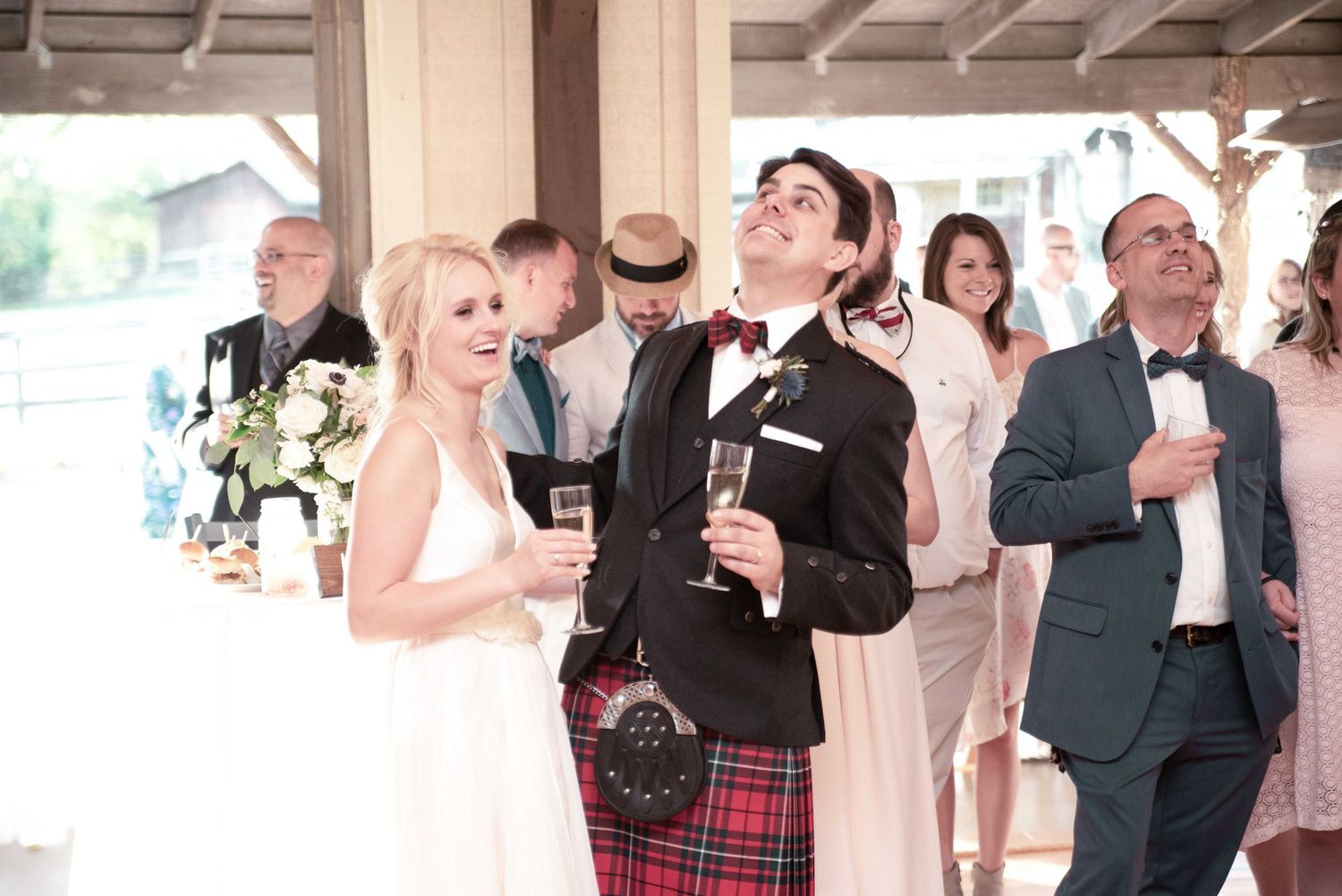 Bride and groom in Scottish kilt toast with champagne at wedding reception surrounded by guests.