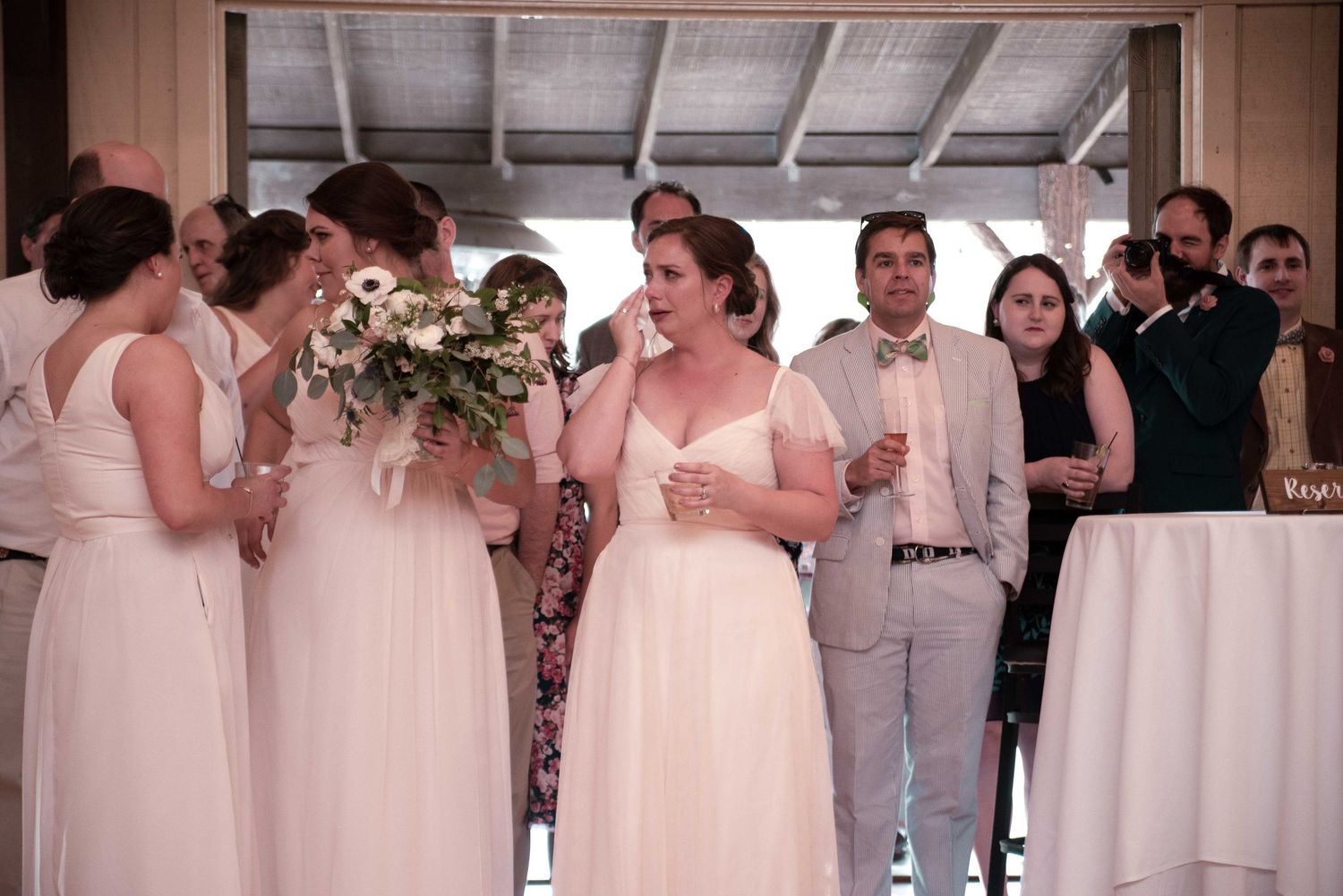 Bridesmaids in white dresses stand together at a wedding reception, one holding a floral bouquet, guests visible in background.