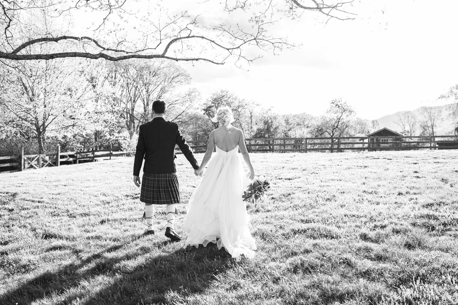 Black and white photo of a bride and groom in a kilt walking hand in hand across a grassy field.
