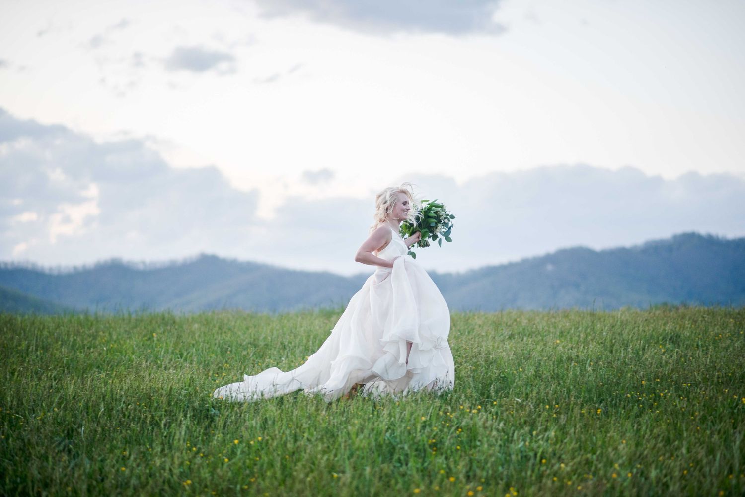 Bride in flowing white gown sitting in green field holding bouquet with mountains behind.