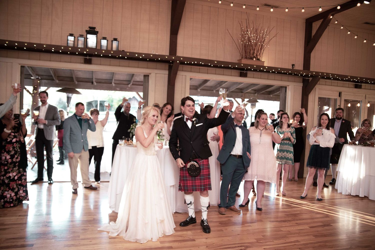 Bride in white gown and groom in red tartan kilt celebrate wedding reception entrance on dance floor with cheering guests.